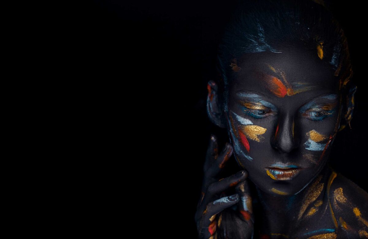 A person with dark, artistic body paint highlighted with vibrant orange and gold patterns, posing thoughtfully against a black background.