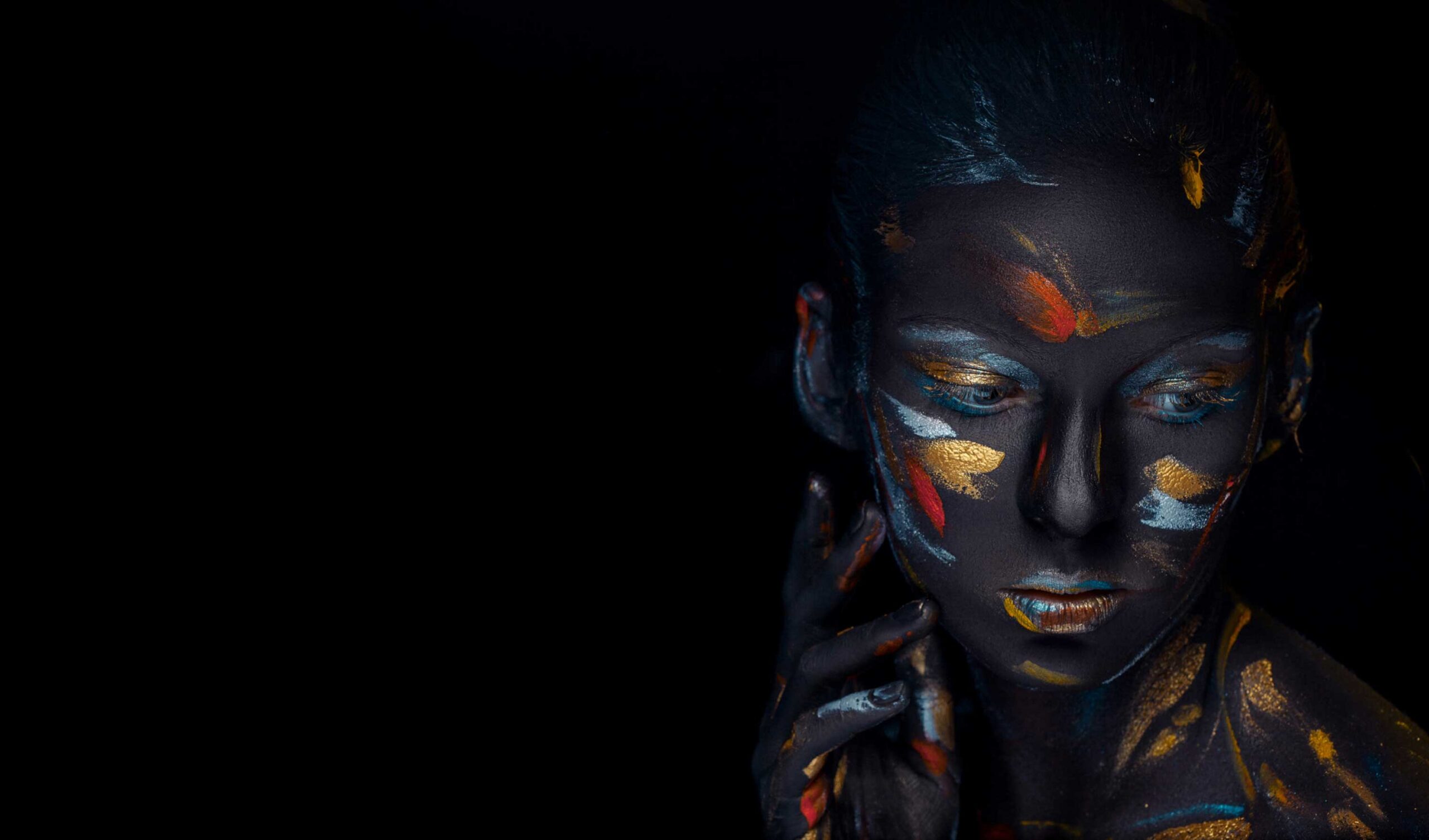 A person with dark, artistic body paint highlighted with vibrant orange and gold patterns, posing thoughtfully against a black background.