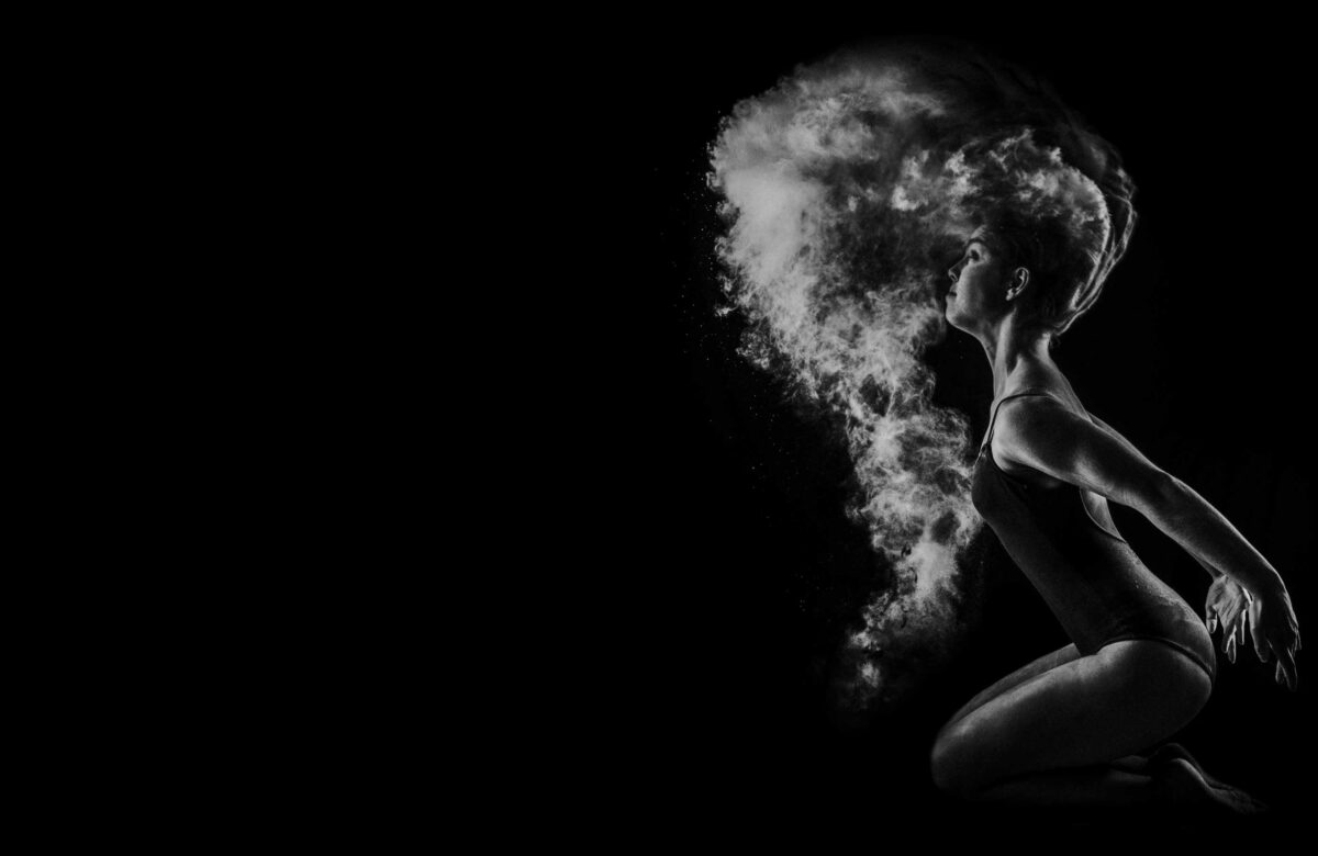A monochrome image of a woman kneeling and dramatically exhaling a cloud of fine powder, creating a striking contrast against the dark background.