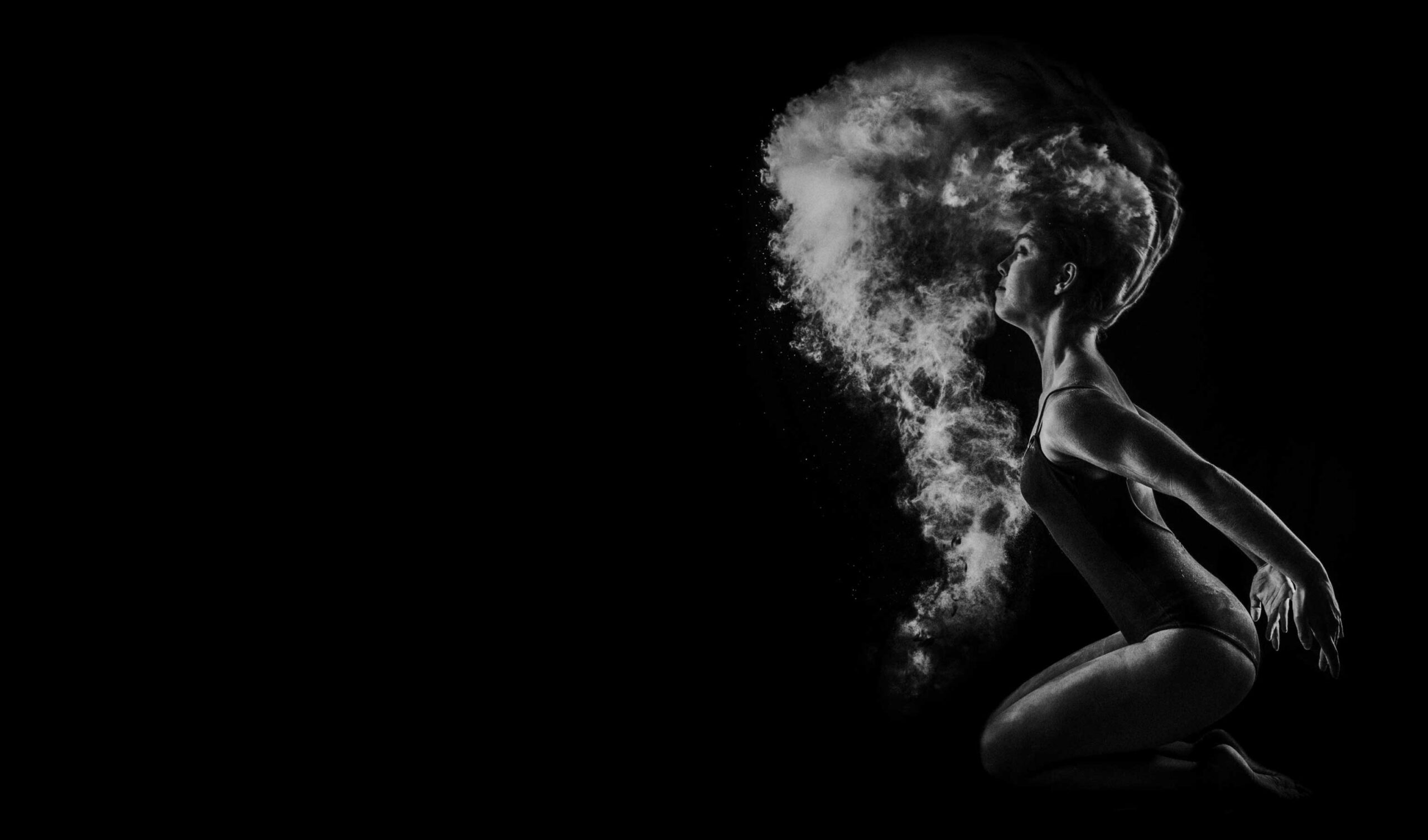 A monochrome image of a woman kneeling and dramatically exhaling a cloud of fine powder, creating a striking contrast against the dark background.