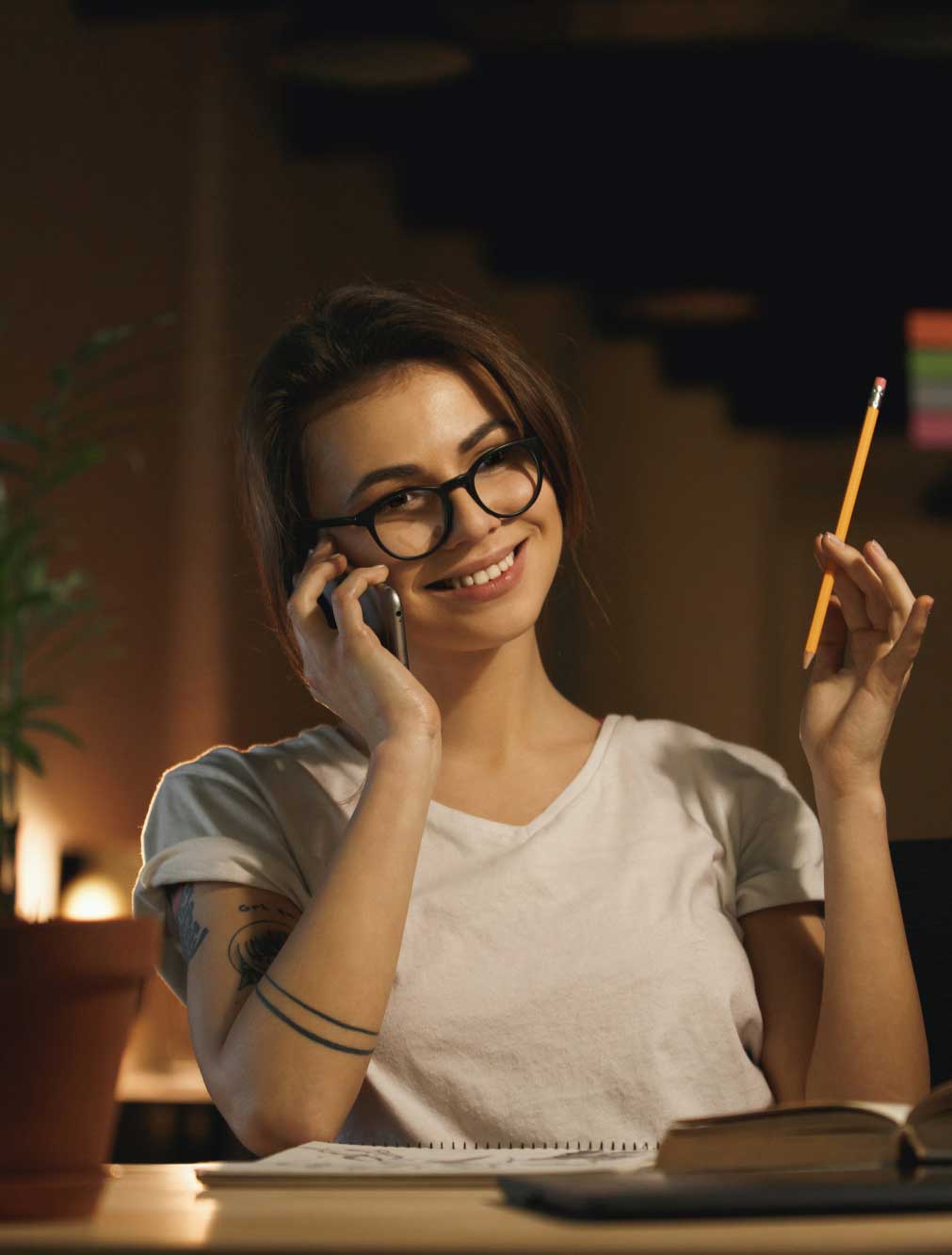A smiling woman with glasses, holding a pencil and talking on the phone, sits by her desk illuminated by a lamp in a dimly lit room.