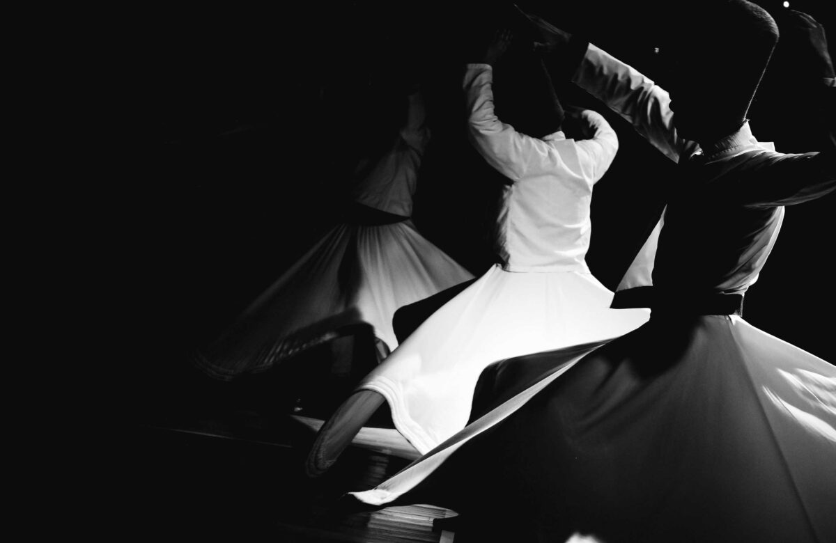 Black and white image of two dancers in motion, wearing flowing skirts and stylish hats, captured in a dynamic, twirling pose on a dark background.