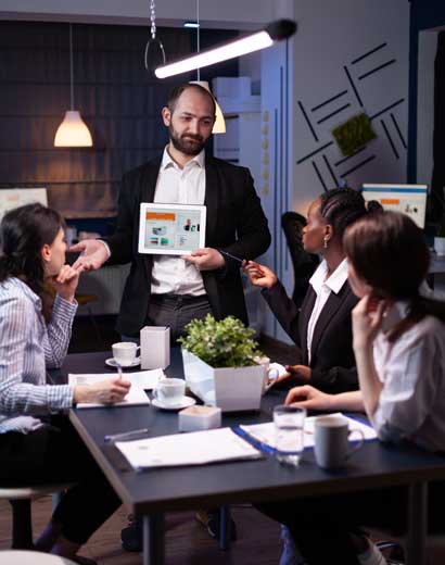 A business presentation in a modern office: A man stands holding a tablet showing graphs, engaging with seated colleagues who are listening attentively.