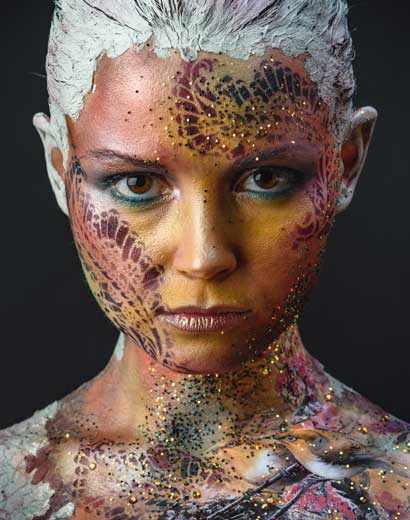 A woman with artistic makeup featuring white textures and colorful patterns on her face and shoulders, against a dark background.