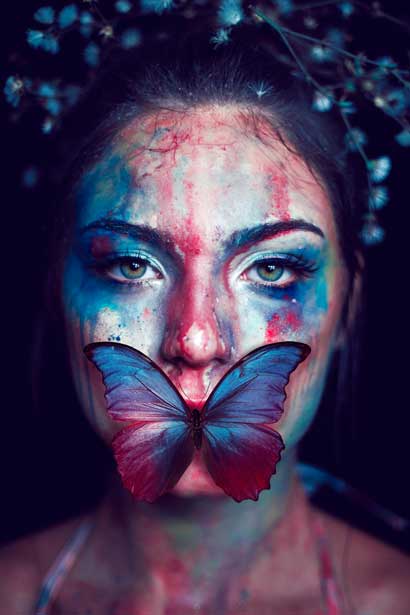 A woman with artistic makeup in blue and red hues, a butterfly covering her mouth, and flowers in her hair against a dark background.