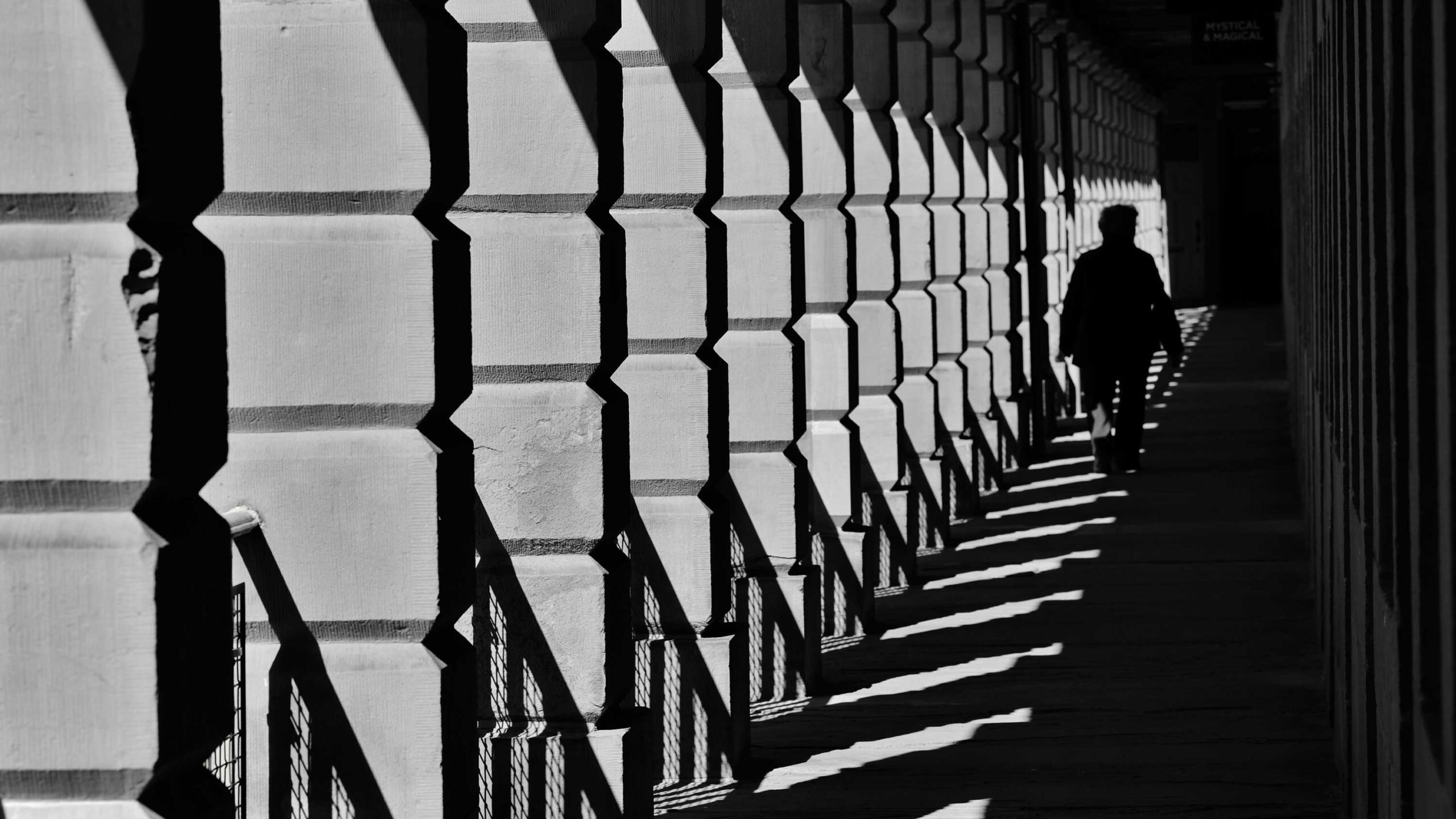 Black and white image of a person walking through a corridor with a patterned series of shadowed archways creating a dramatic play of light and shadow.