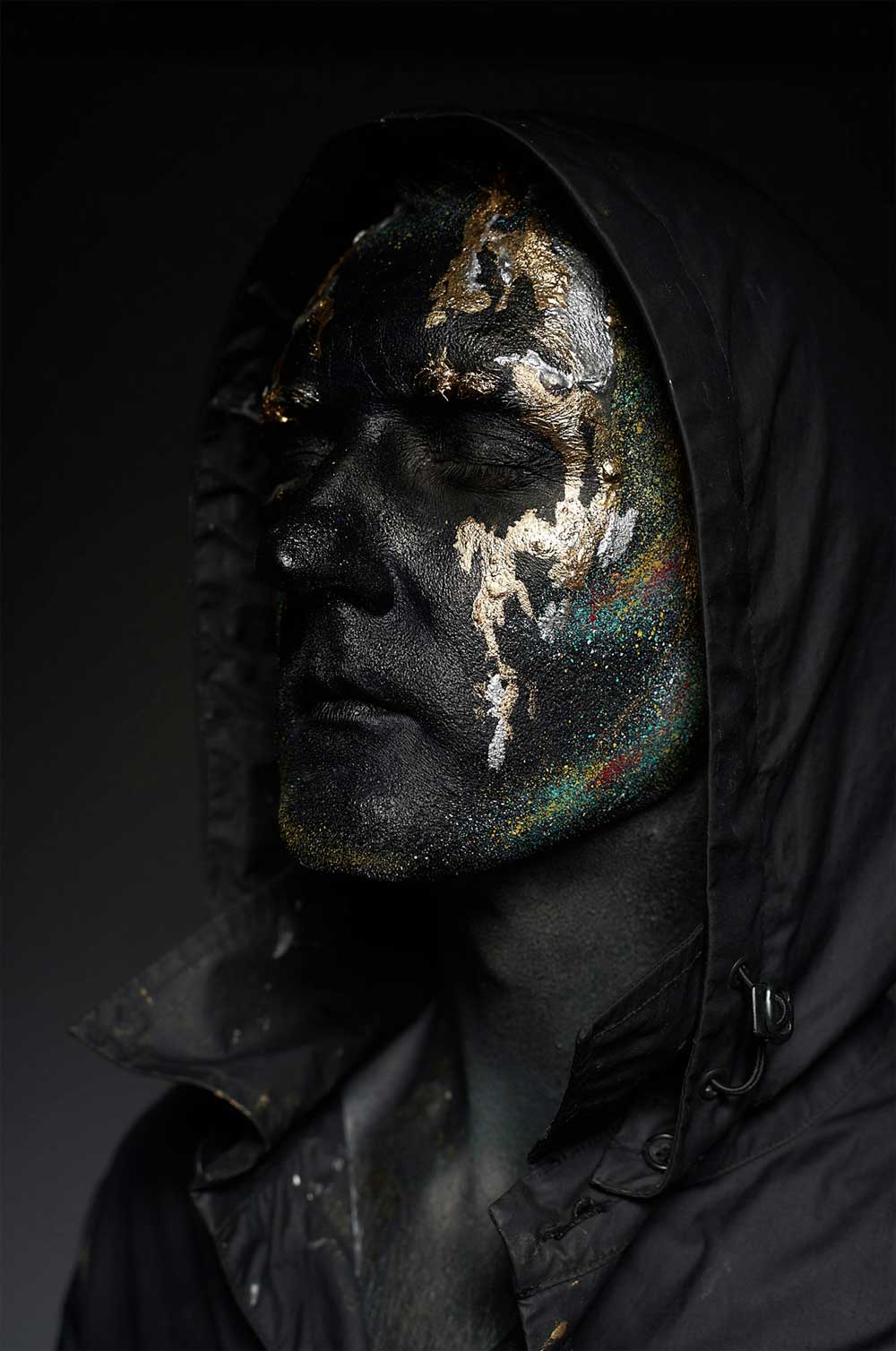 a portrait of a man wearing a black hoodie and his face is also black having an abstract gold design on his face with a dark and black background.