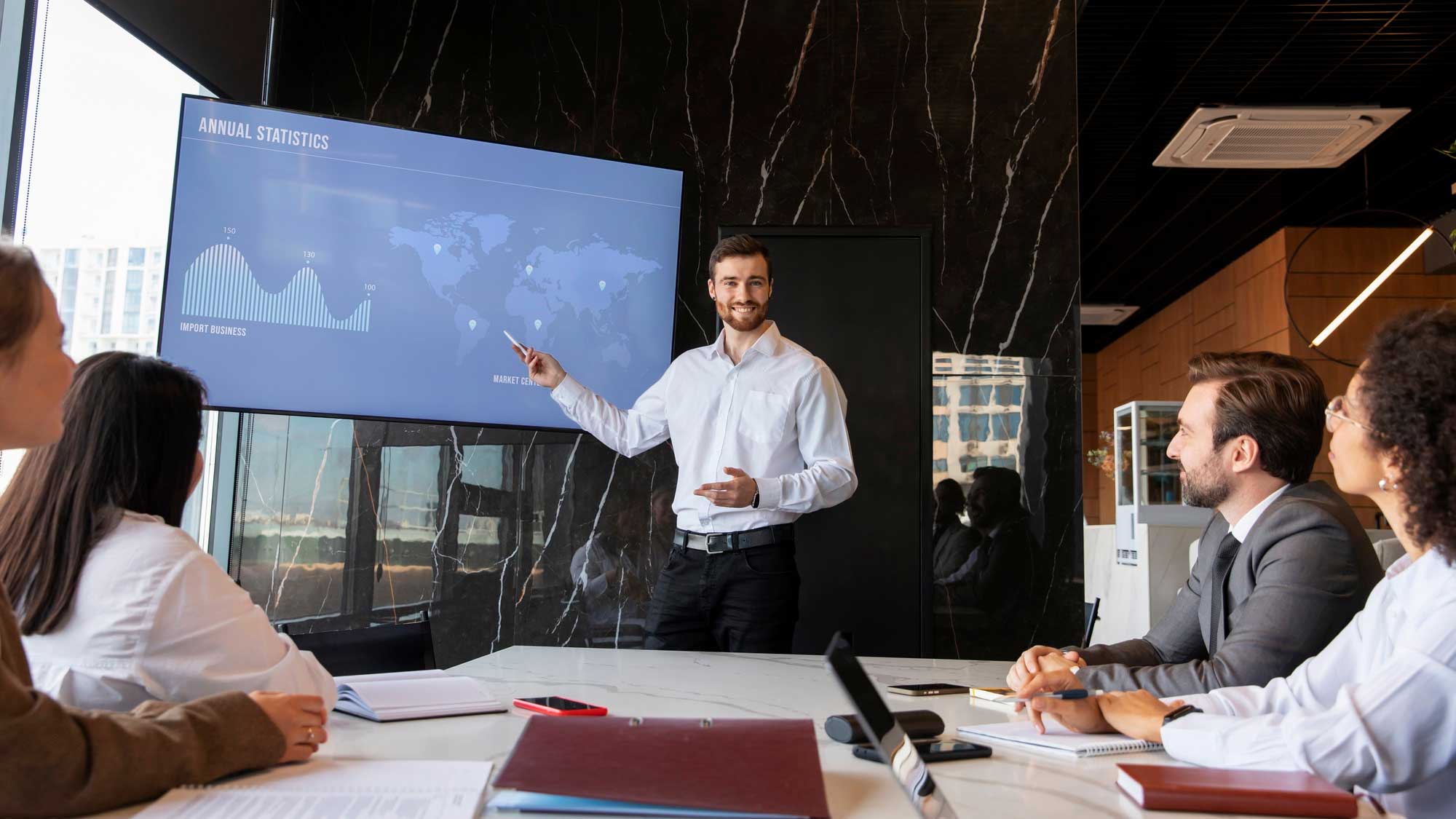 A man presents annual statistics on a screen to colleagues in a meeting room. the screen displays graphs and a world map. three attentive listeners sit at the table, which has documents and laptops.