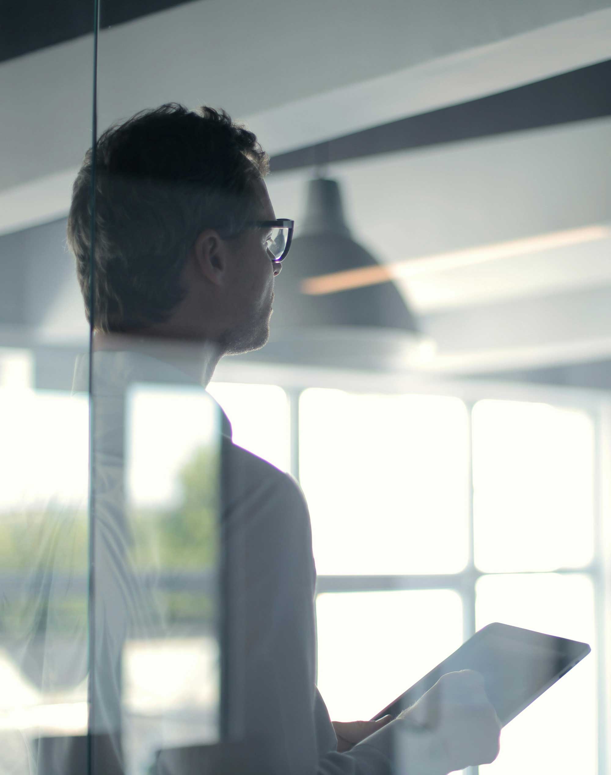 A businessman with glasses holding a tablet, gazing out of a sunlit office.