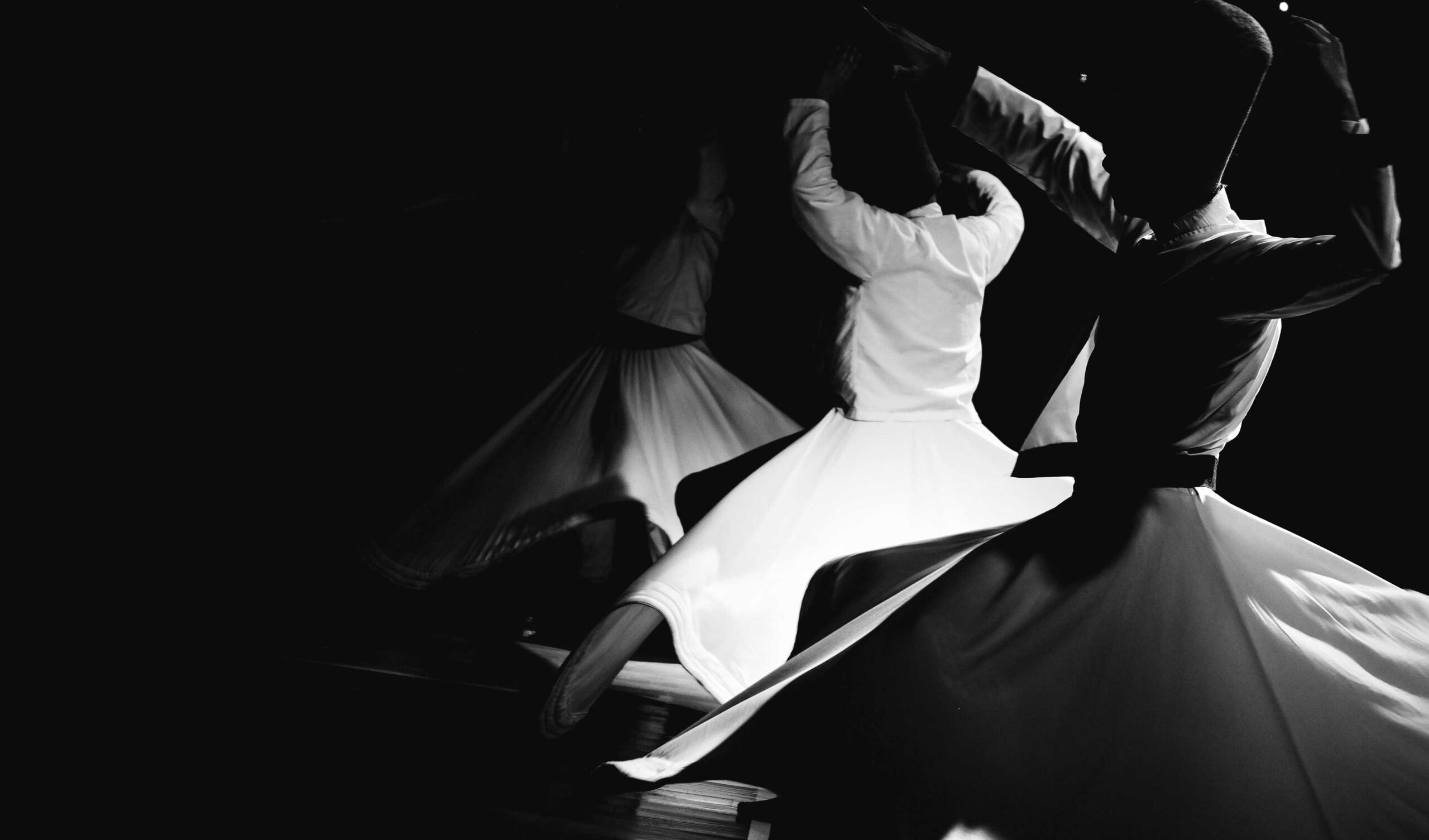 Black and white image of two dancers in motion, wearing flowing skirts and stylish hats, captured in a dynamic, twirling pose on a dark background.