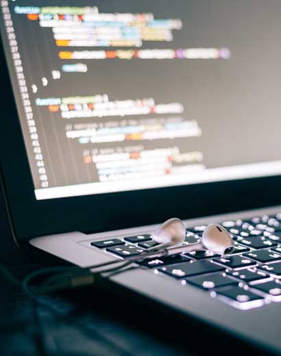 A close-up photo of a laptop keyboard illuminated by the screen displaying colorful lines of code, with white earbuds resting on the keyboard.