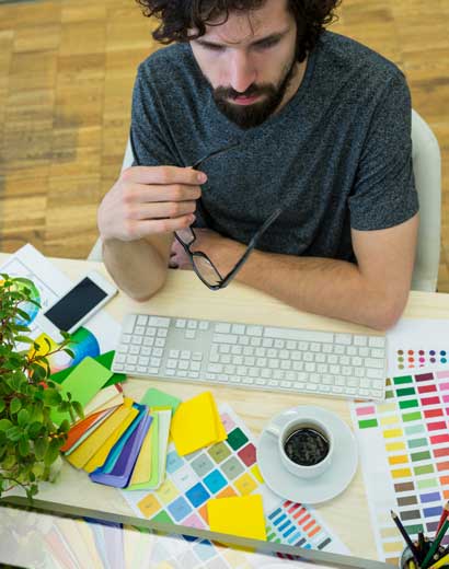 A man sitting at a desk, studying various color swatches beside a keyboard, smartphone, and a cup of coffee, holding his glasses and focusing intently.