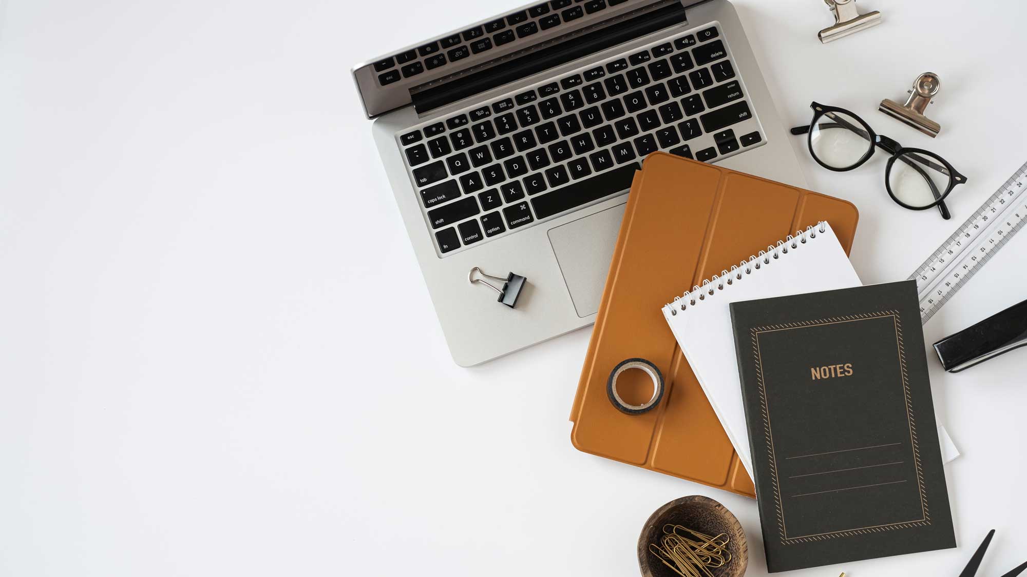 A neatly organized workspace with a laptop, mechanical keyboard, coffee cup, and stationery items on a white surface.