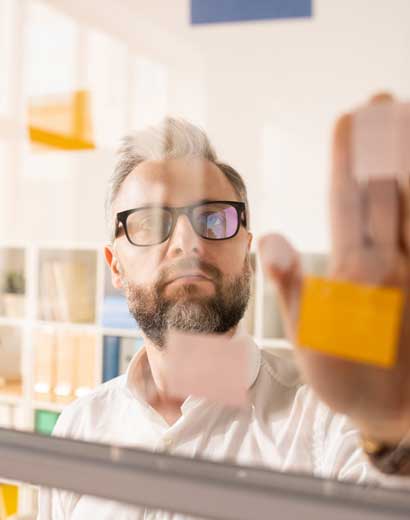 A man with glasses and a beard placing a sticky note on a glass wall, viewed through the glass in an office environment.