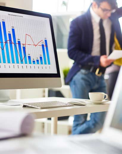 A business professional examines documents in an office with a coffee cup, notebook, and a computer screen displaying colorful charts and graphs in the foreground.